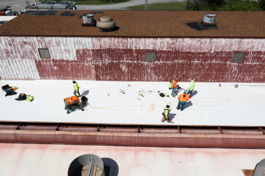 A group of workers in safety gear install roofing material on a flat commercial roof, using equipment and tools spread across the white surface.