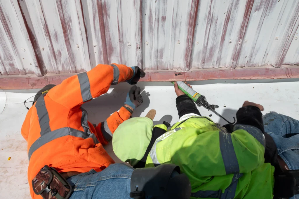 Two workers in safety gear apply sealant to the base of a metal wall, working closely on a construction or maintenance task.