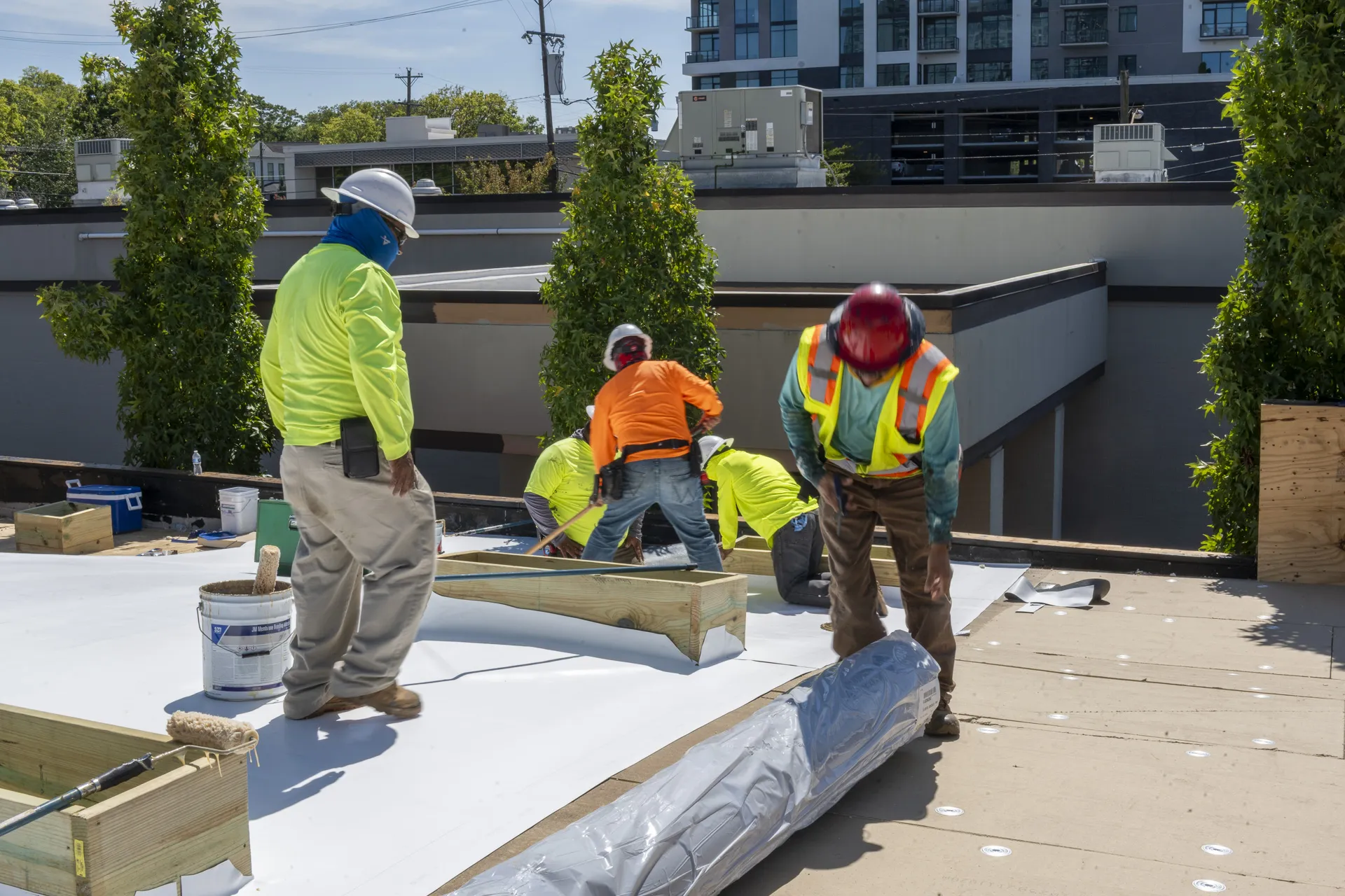 Four construction workers in safety gear install roofing materials on a flat building roof, with tools and equipment scattered around. Trees and buildings are visible in the background.